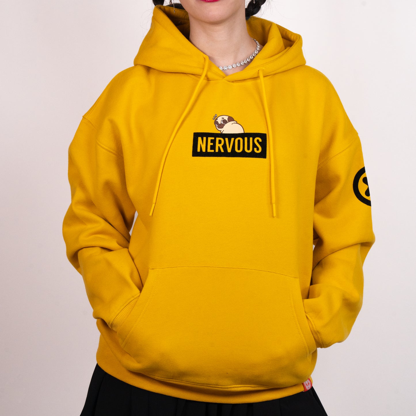 A person wearing a yellow hoodie with a graphic of a scared-looking pug above a black rectangle with the yellow text "NERVOUS". The graphic is in the centre of the chest of the hoodie. There is a black X with a black circle around it on the top-left of one of the the sleeves. The model has their hands in the hoodie front pocket. There is a small red tag on the bottom hem of the hoodie.