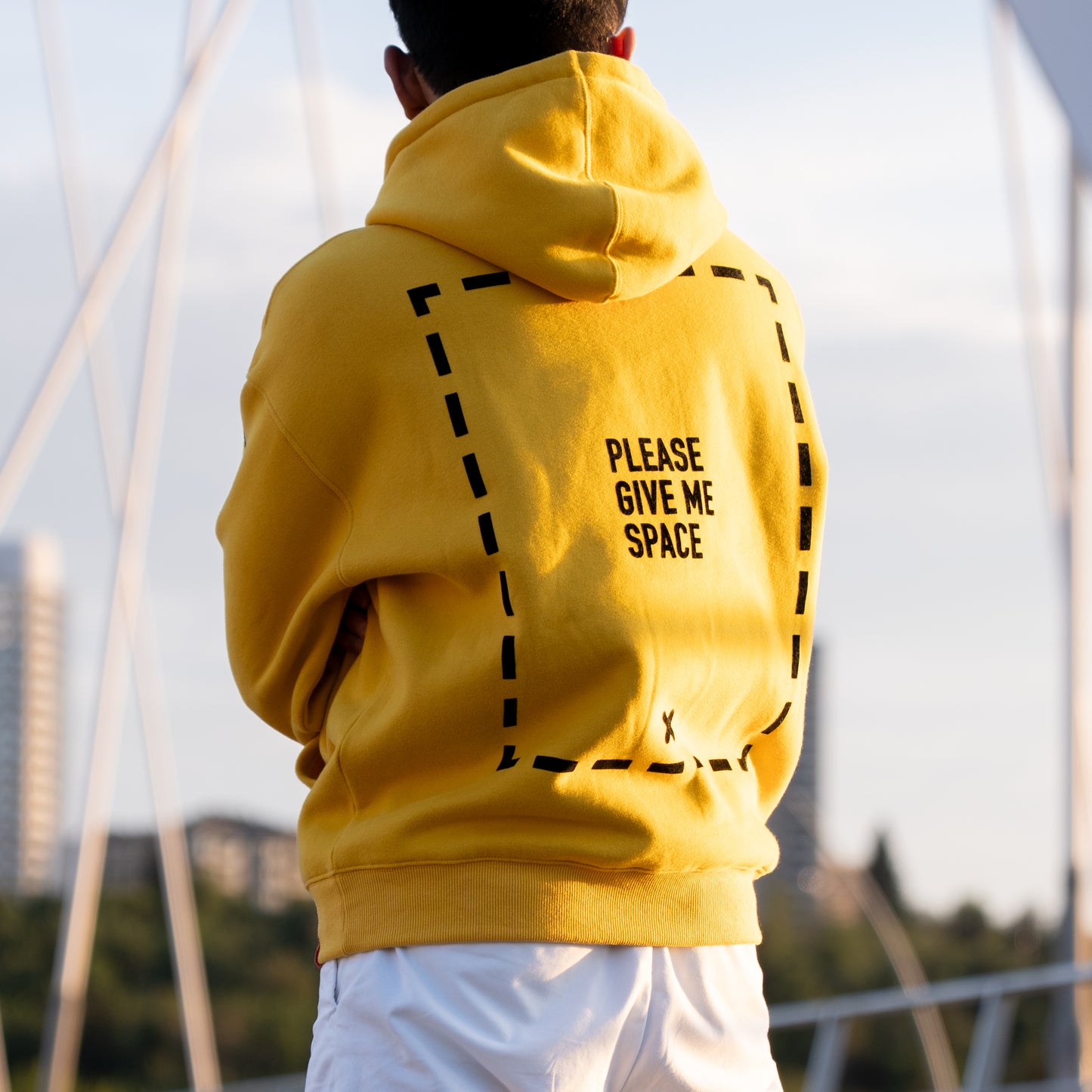 A person facing away from the camera wearing a yellow hoodie with a graphic of a dotted line rectangle covering the majority of the back of the garment. In the center of the rectangle is the text "PLEASE GIVE ME SPACE". The graphic is in the centre of the back of the hoodie. The model appears to be folding their arms and looking out at an out-of-focus cityscape. They are wearing white pants.