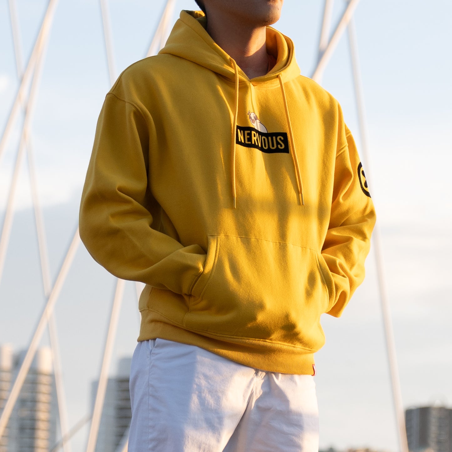 A person standing 3/4 towards the camera wearing a yellow hoodie with a graphic of a scared-looking pug above a black rectangle with the yellow text "NERVOUS". The graphic is in the centre of the chest of the hoodie. There is a black X with a black circle around it on the top-left of one of the the sleeves. The model has their hands in the hoodie front pocket. They are wearing white pants. The background is an out-of-focus cityscape.