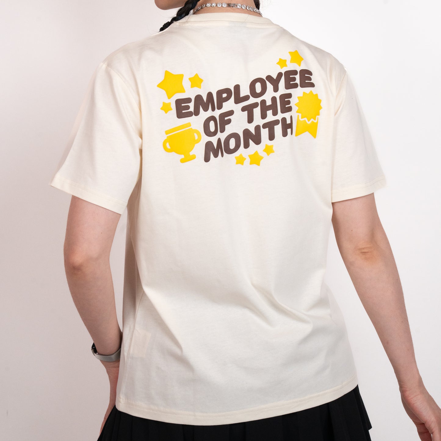Person facing away from the camera, wearing a cream-coloured t-shirt with a graphic of all-caps brown text that says 'EMPLOYEE OF THE MONTH'. The text is surrounded by yellow images of multiple sized stars, a trophy, and an award ribbon. The graphic is in the upper center of the back of the t-shirt.