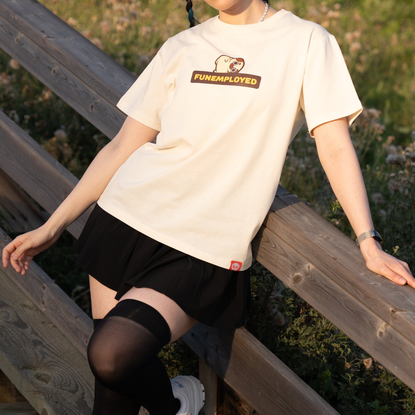 Person wearing a cream-coloured t-shirt with a graphic of Puglie laying down on the bolded all-caps yellow text outlined in black that says 'FUNEMPLOYED'. The graphic is in the center of the chest of the t-shirt.