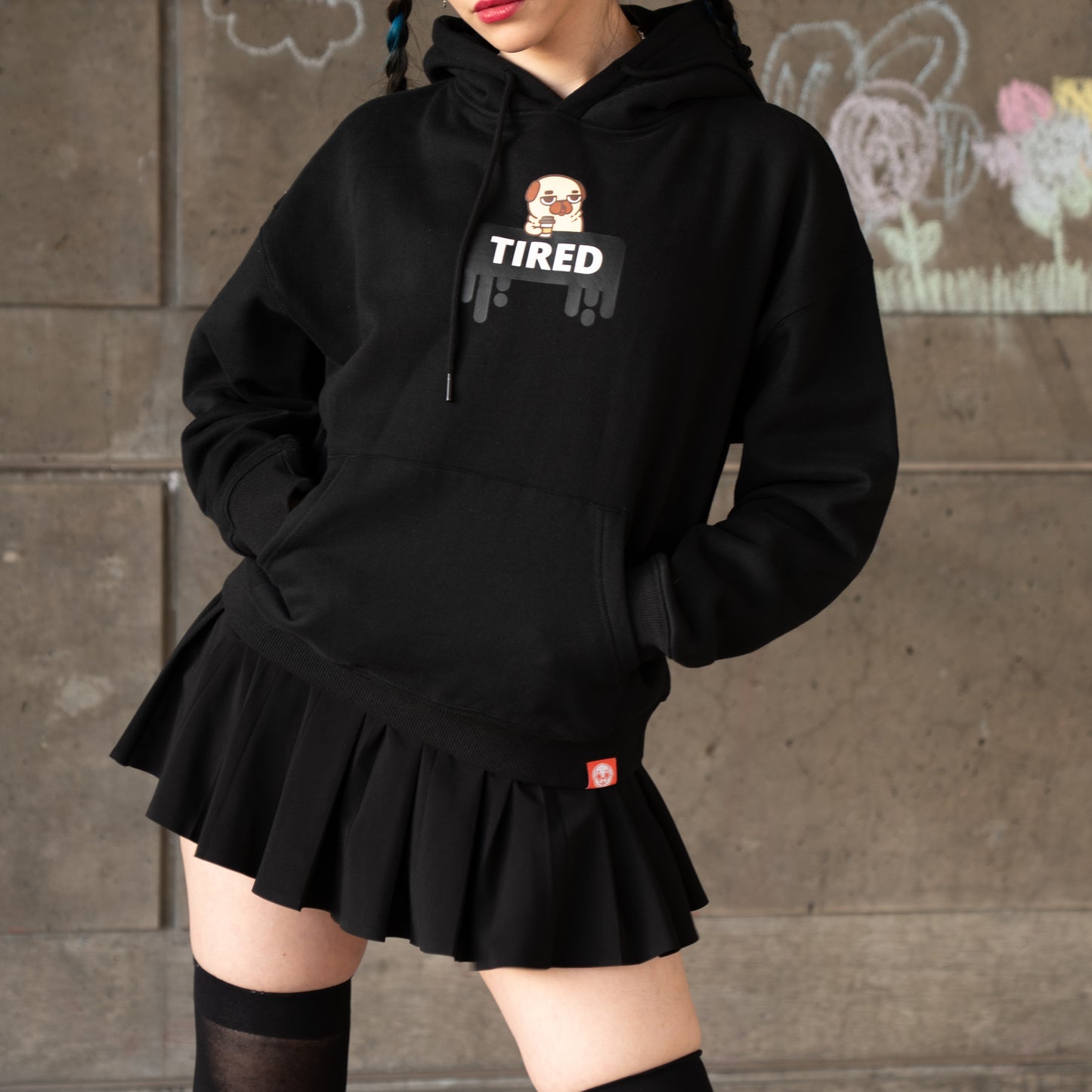 A person wearing a black hoodie with a graphic of a tired-looking pug holding a to-go coffee cup above a black dripping rectangle with the white text "TIRED". The graphic is in the centre of the chest of the hoodie. There is a small red tag on the bottom hem of the hoodie. The model has their hands in the hoodie front pocket with their hip cocked to the side, and is wearing a black skirt and thigh-high stockings.