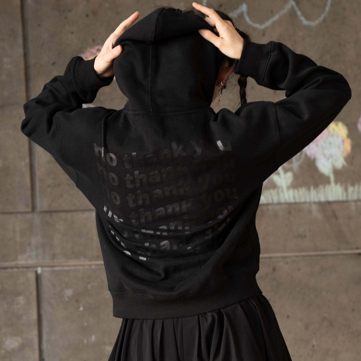 A person facing away from the camera wearing a black hoodie with a graphic of wavy repeated text on the back that says "no thank you". The graphic is in the centre of the back of the hoodie. There is an off-black "X" graphic just below the text. The model has their arms up grasping the pulled up hood. They are wearing a black skirt.