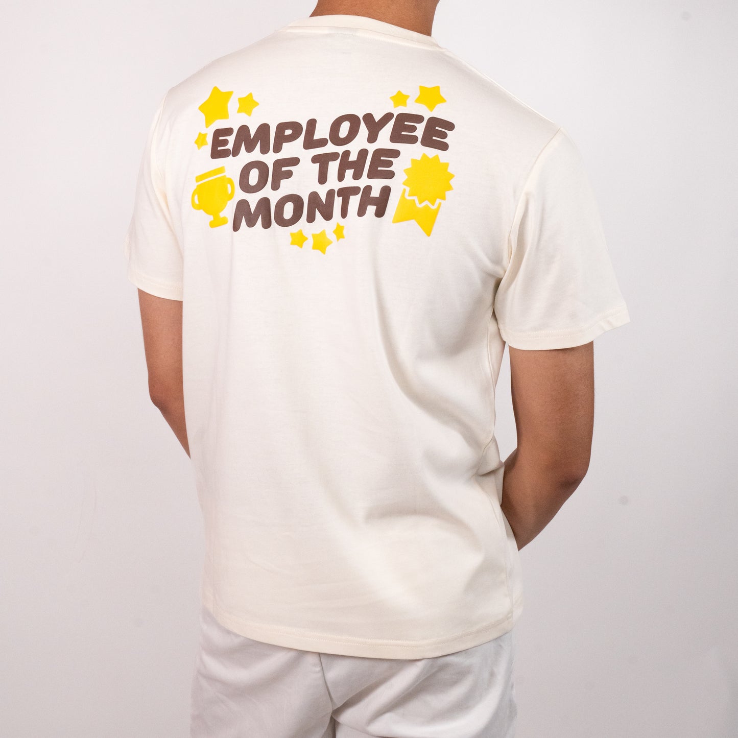 Person facing away from the camera, wearing a cream-coloured t-shirt with a graphic of all-caps brown text that says 'EMPLOYEE OF THE MONTH'. The text is surrounded by yellow images of multiple sized stars, a trophy, and an award ribbon. The graphic is in the upper center of the back of the t-shirt.