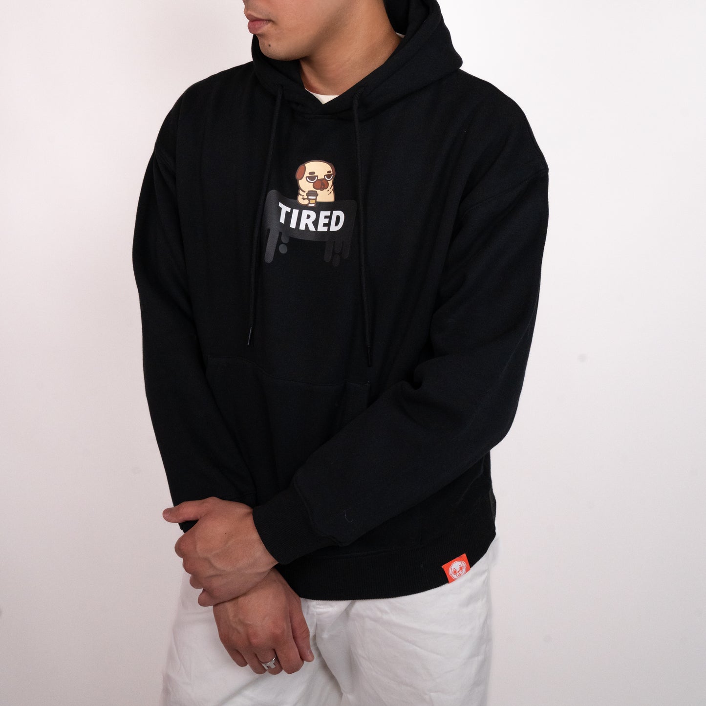 A person wearing a black hoodie with a graphic of a tired-looking pug holding a to-go coffee cup above a black dripping rectangle with the white text "TIRED". The graphic is in the centre of the chest of the hoodie. There is a small red tag on the bottom hem of the hoodie. The model has their left hand clasped over their right wrist. They are wearing white pants.