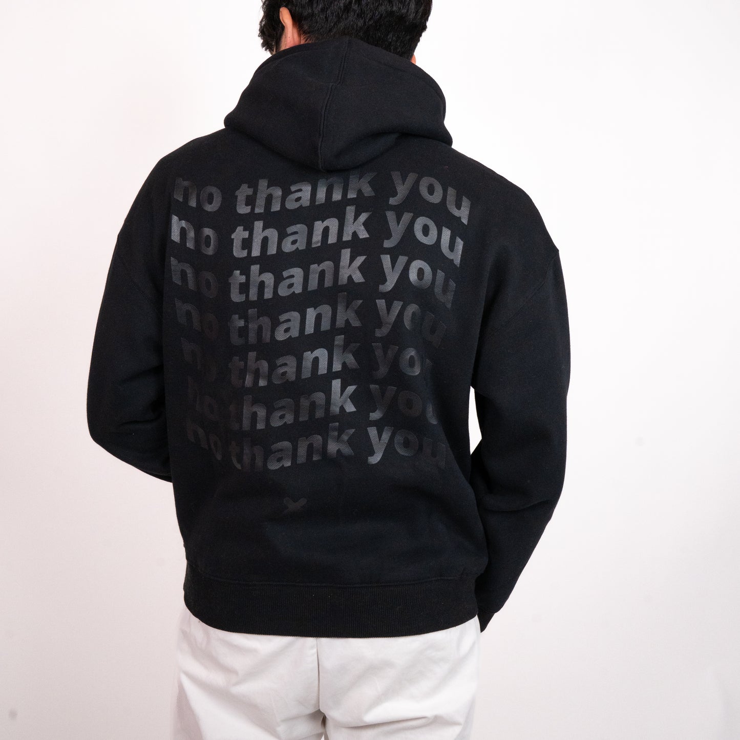 A person facing away from the camera wearing a black hoodie with a graphic of wavy repeated text on the back that says "no thank you". The graphic is in the centre of the back of the hoodie. There is an off-black "X" graphic just below the text. The model has relaxed arms with one slightly raised, and is wearing white pants.