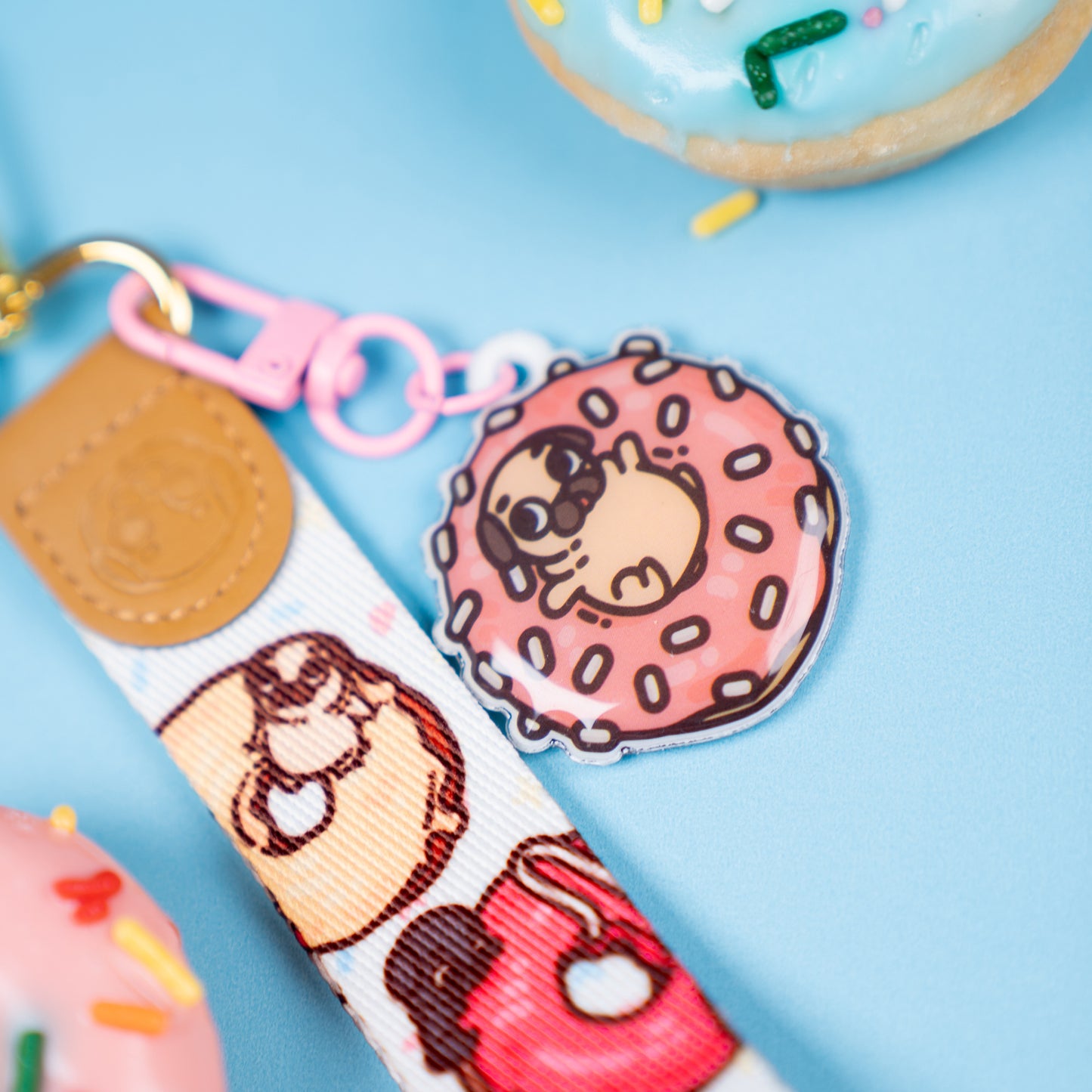 Keychain with a wrist strap featuring a design of various doughnuts with tiny pugs playing on them, with a leather-looking clasp embossed with a pug face. The leather-looking clasp is stitched to the wristlet and attached to a gold oval hoop. In focus is a pink hook clasp handing from the gold hoop with a cute charm featuring Puglie sitting in a pink doughnut with white sprinkles. The background is blue with doughnuts.