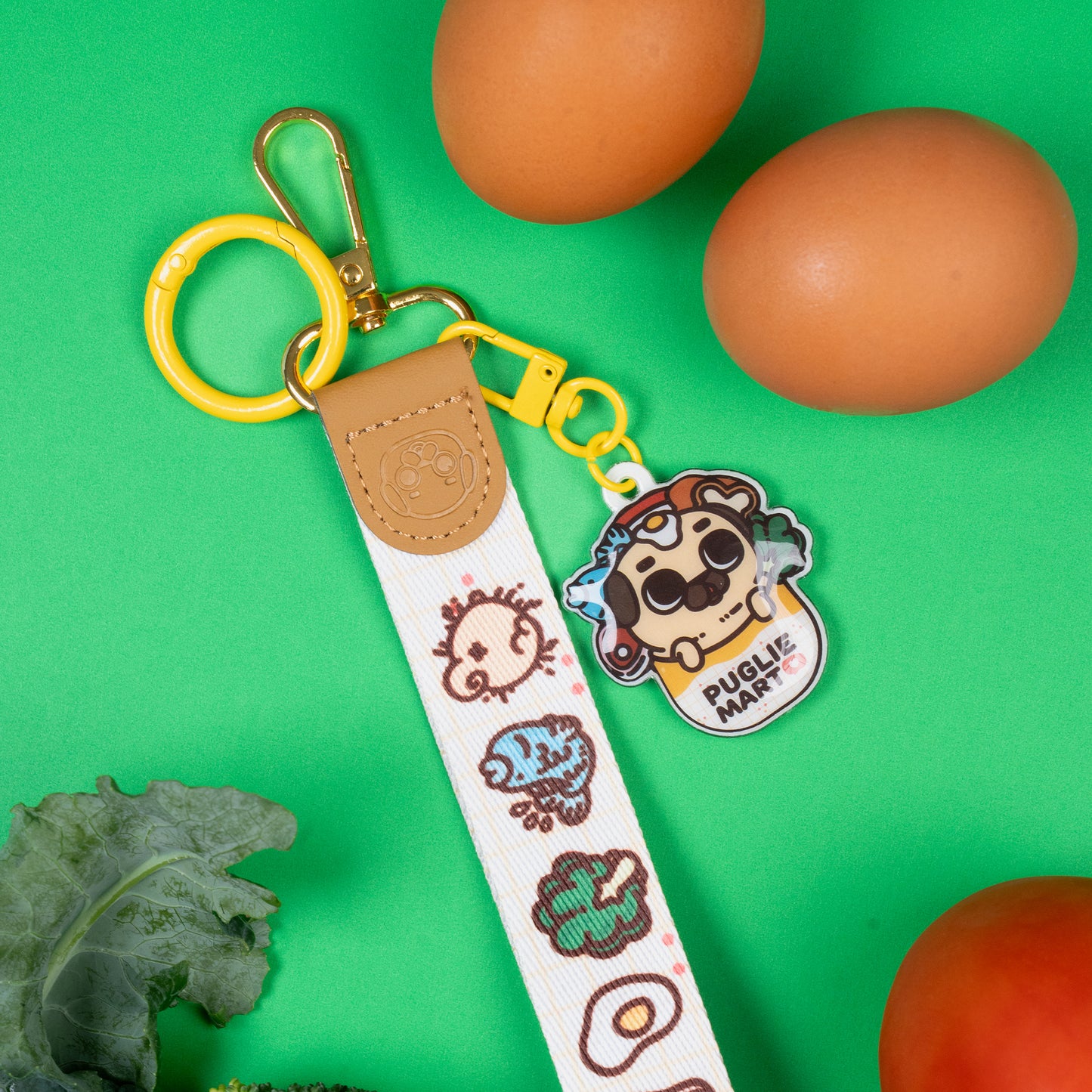 Keychain with a wrist strap featuring a design of many food items and one pug butt, with a leather-looking clasp embossed with a pug face. The leather-looking clasp is stitched to the wristlet and attached to a gold oval hoop, which has a yellow jump ring, a lobster clasp, and a hook clasp attached with a cute charm featuring Puglie in a bag with a bunch of food items surrounding him. The bag says "PUGLIE MART ❤️". The keychain is displayed on a green background surrounded by grocery items.