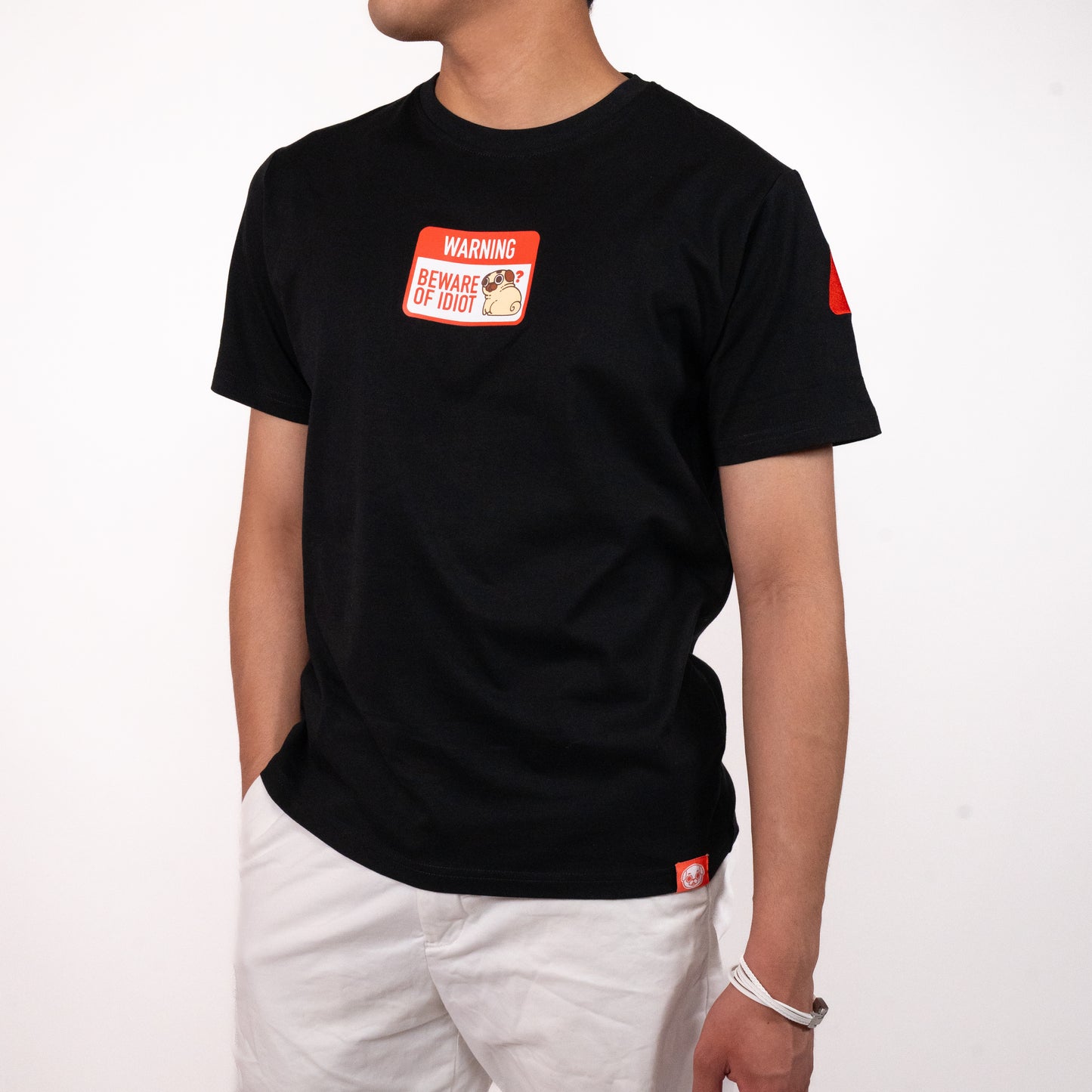 Person wearing a black t-shirt with a graphic of a warning sign that says 'WARNING BEWARE OF PUGLIE' with an image of Puglie sitting down and looking confused. The graphic is in the center of the chest of the t-shirt. A small red design is visible on the shoulder and on the bottom hem of the shirt. The model is posing with one hand in their hand in their pocket and is wearing a pair of white pants.