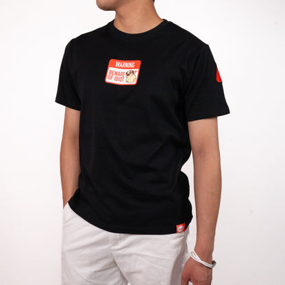 Person wearing a black t-shirt with a graphic of a warning sign that says 'WARNING BEWARE OF PUGLIE' with an image of Puglie sitting down and looking confused. The graphic is in the center of the chest of the t-shirt. A small red design is visible on the shoulder and on the bottom hem of the shirt. The model is posing with one hand in their hand in their pocket and is wearing a pair of white pants.