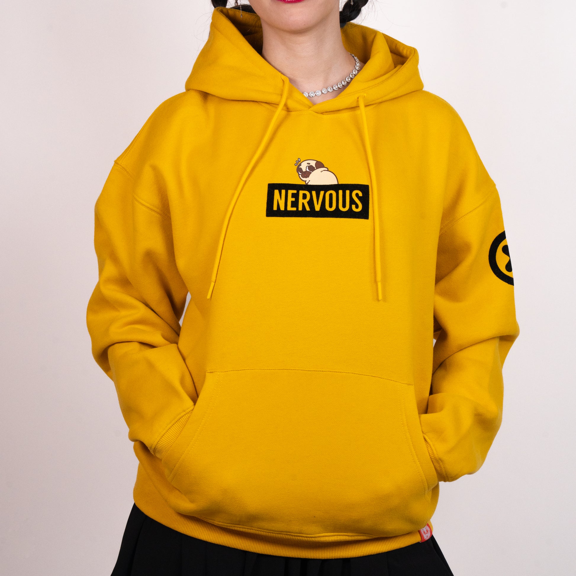 A person wearing a yellow hoodie with a graphic of a scared-looking pug above a black rectangle with the yellow text "NERVOUS". The graphic is in the centre of the chest of the hoodie. There is a black X with a black circle around it on the top-left of one of the the sleeves. The model has their hands in the hoodie front pocket. There is a small red tag on the bottom hem of the hoodie.