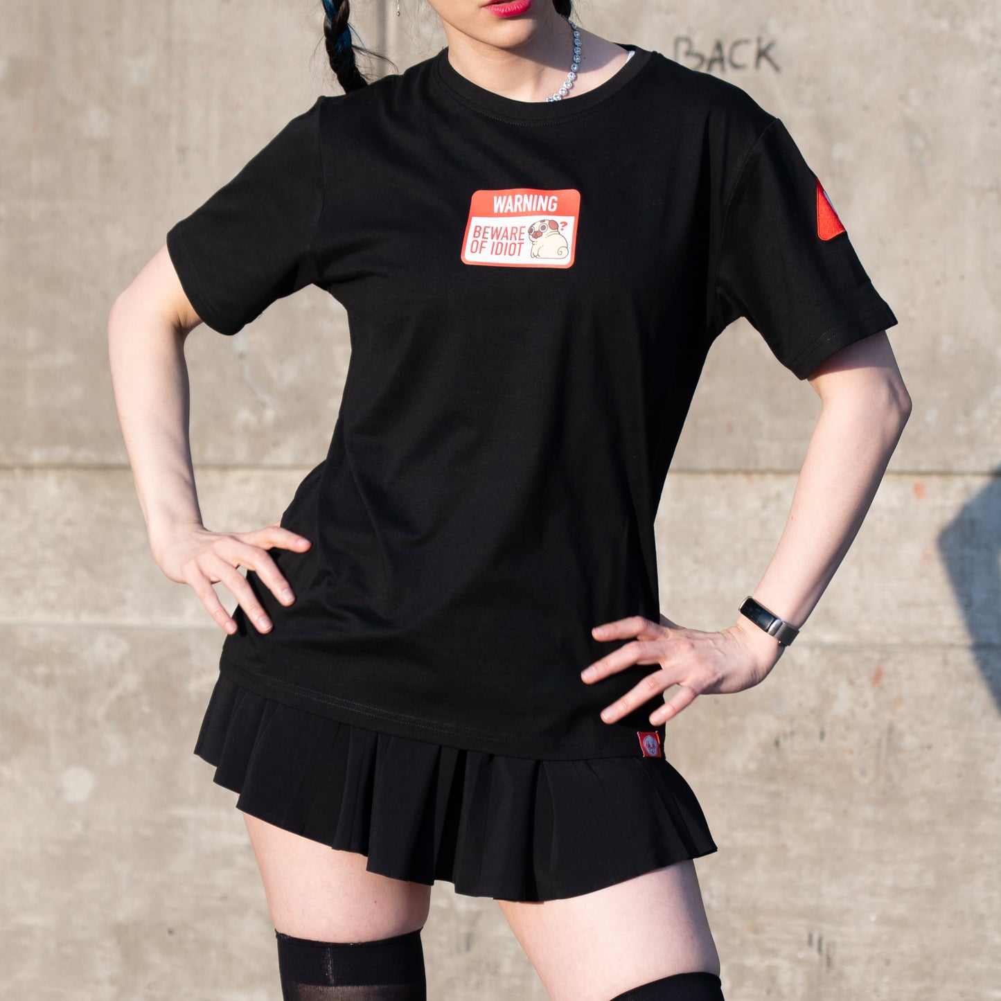 Person wearing a black t-shirt with a graphic of a warning sign that says 'WARNING BEWARE OF PUGLIE' with an image of Puglie sitting down and looking confused. The graphic is in the center of the chest of the t-shirt. A small red design is visible on the shoulder and on the bottom hem of the shirt. The model is posing with their hands on their hips. They have a femme style and are wearing a short black skirt and thigh-high stockings.