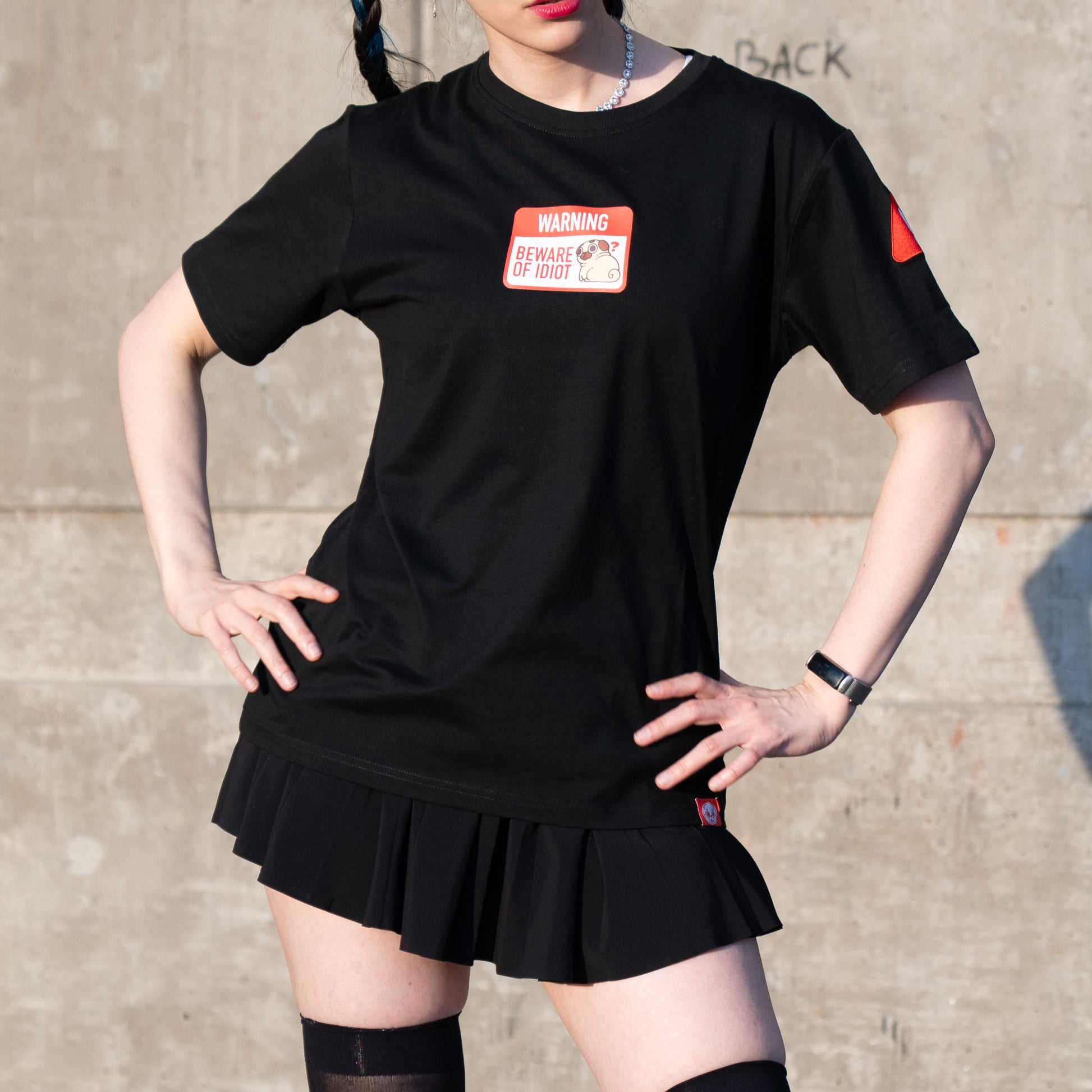 Person wearing a black t-shirt with a graphic of a warning sign that says 'WARNING BEWARE OF PUGLIE' with an image of Puglie sitting down and looking confused. The graphic is in the center of the chest of the t-shirt. A small red design is visible on the shoulder and on the bottom hem of the shirt. The model is posing with their hands on their hips. They have a femme style and are wearing a short black skirt and thigh-high stockings.