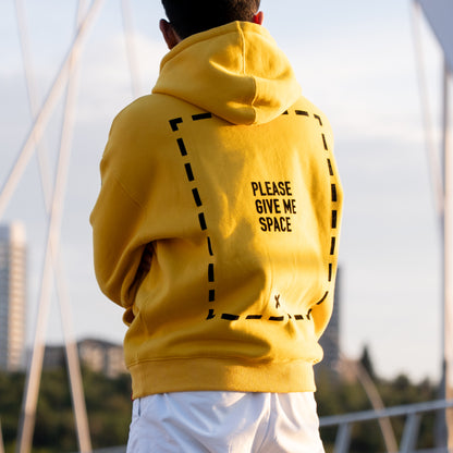A person facing away from the camera wearing a yellow hoodie with a graphic of a dotted line rectangle covering the majority of the back of the garment. In the center of the rectangle is the text "PLEASE GIVE ME SPACE". The graphic is in the centre of the back of the hoodie. The model appears to be folding their arms and looking out at an out-of-focus cityscape. They are wearing white pants.