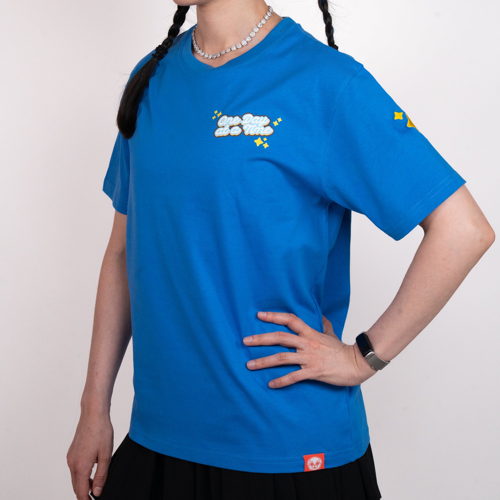 Person wearing a blue t-shirt with a graphic of light blue and white cursive text saying 'One Day at a Time". The text is surrounded by yellow sparkle emoji. The graphic is in the top-left of the chest of the t-shirt. A small yellow design is visible on the shoulder. A small red tag with a Puglie face on the bottom hem of the shirt. The model is posing with one hand on their hip.