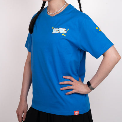 Person wearing a blue t-shirt with a graphic of light blue and white cursive text saying 'One Day at a Time". The text is surrounded by yellow sparkle emoji. The graphic is in the top-left of the chest of the t-shirt. A small yellow design is visible on the shoulder. A small red tag with a Puglie face on the bottom hem of the shirt. The model is posing with one hand on their hip.