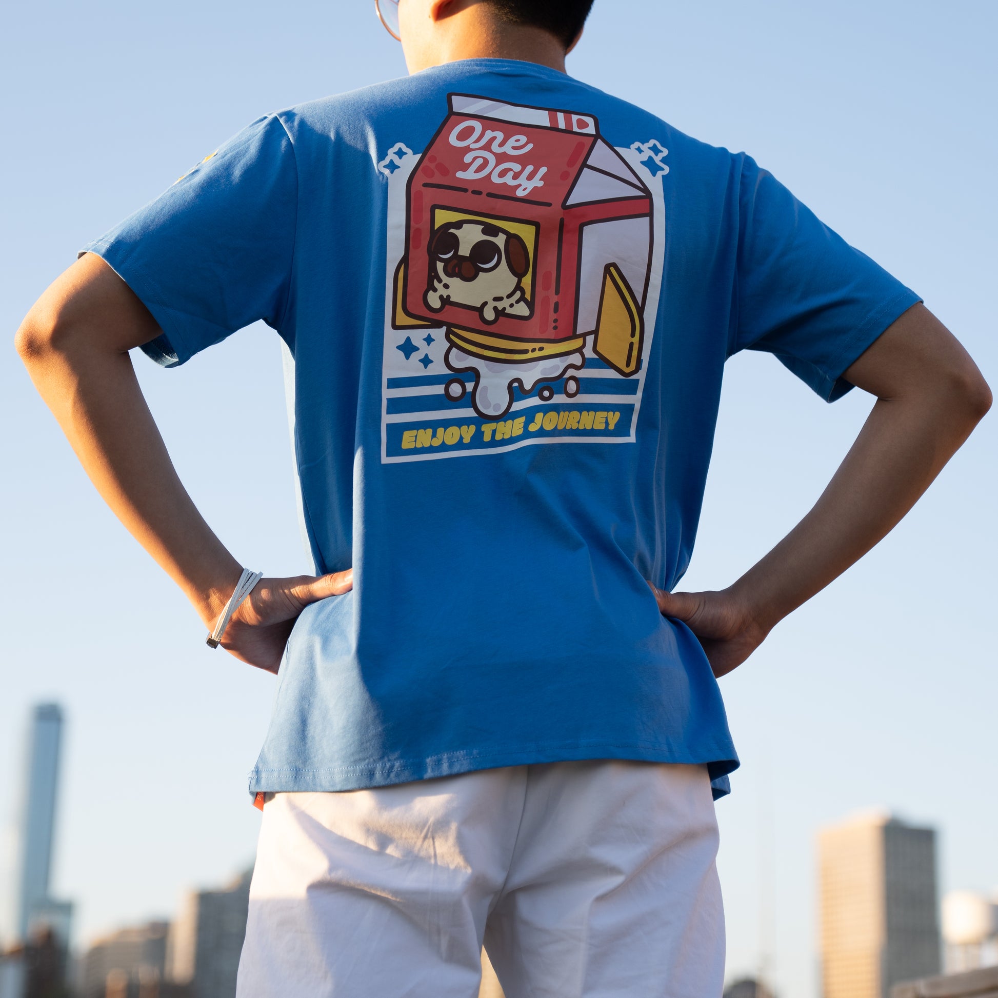 A person facing away from the camera wearing a blue t-shirt with a graphic of a red, white, and yellow rocket ship shaped like a milk carton with a tan-colored pug peeking out of window. The rocket appears to be blasting milk out the bottom, and the graphic is surrounded by a geometric white diagonal rectangle with cut-out lines and sparkle emoji. The graphic is in the centre of the back of the t-shirt. A small yellow design is visible on the shoulder. The model has their hands on their hips.
