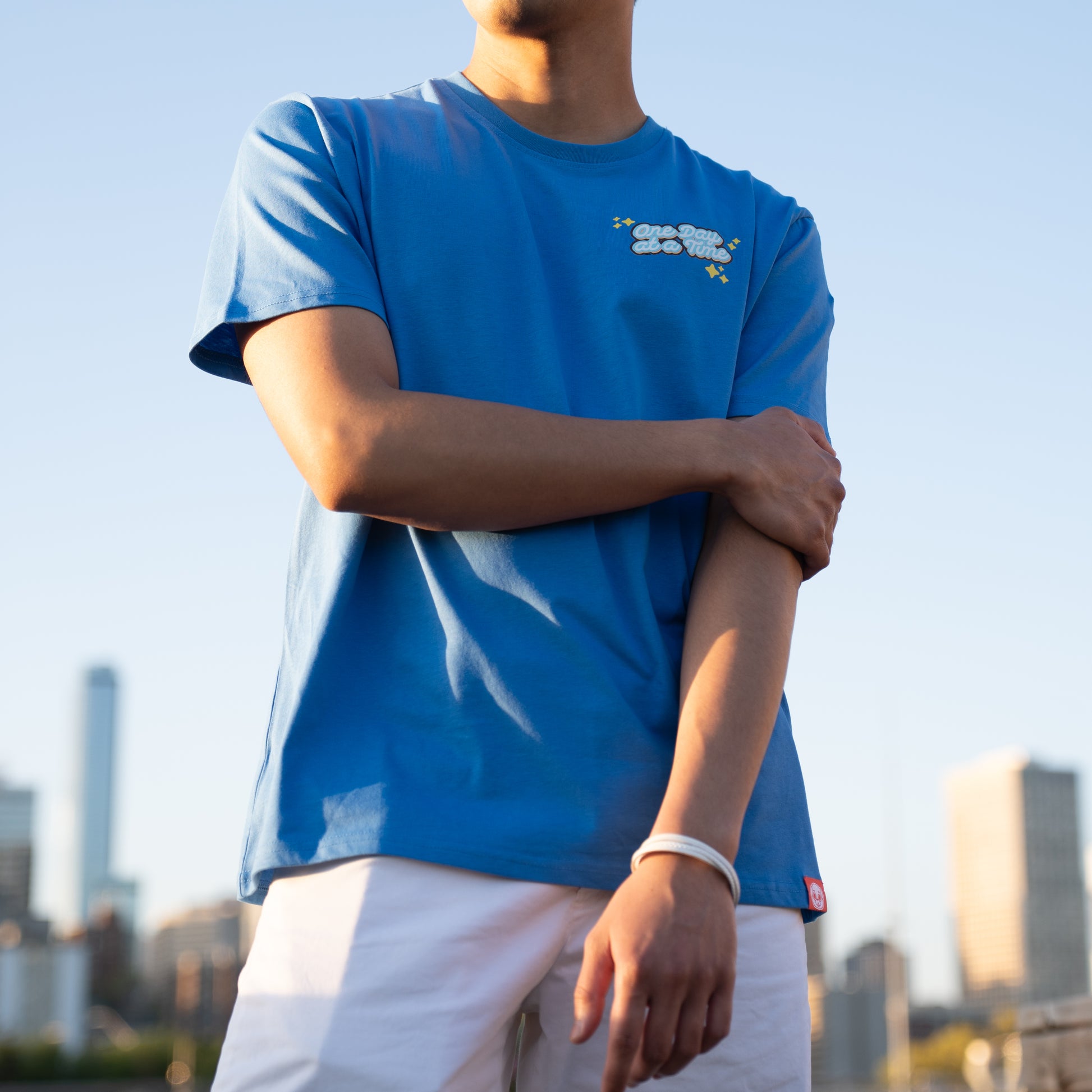 Person wearing a blue t-shirt with a graphic of light blue and white cursive text saying 'One Day at a Time". The text is surrounded by yellow sparkle emoji. The graphic is in the top-left of the chest of the t-shirt. A small red tag with a Puglie face on the bottom hem of the shirt. The model is posing with their right hand grasping their upper left arm. The background is an out of focus cityscape.