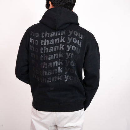 A person facing away from the camera wearing a black hoodie with a graphic of wavy repeated text on the back that says "no thank you". The graphic is in the centre of the back of the hoodie.  There is an off-black "X" graphic just below the text. The model has relaxed arms with one slightly raised, and is wearing white pants.