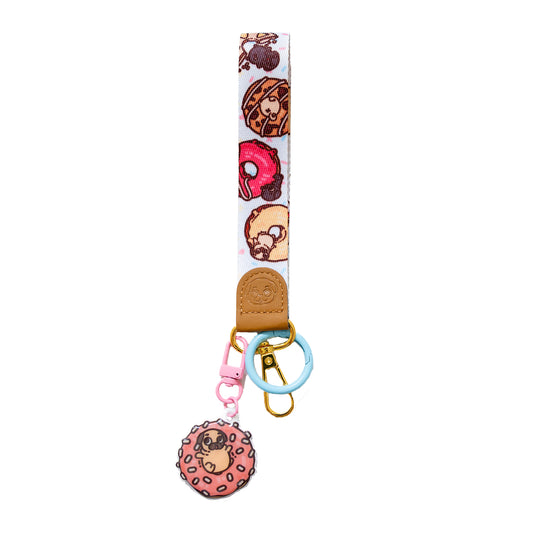 Keychain with a wrist strap featuring a design of various doughnuts with tiny pugs playing on them, with a leather-looking clasp embossed with a pug face. The leather-looking clasp is stitched to the wristlet and attached to a gold oval hoop, which has a light blue jump ring, a gold lobster clasp, and a pink hook clasp attached with a cute charm featuring Puglie sitting in a pink doughnut with white sprinkles.