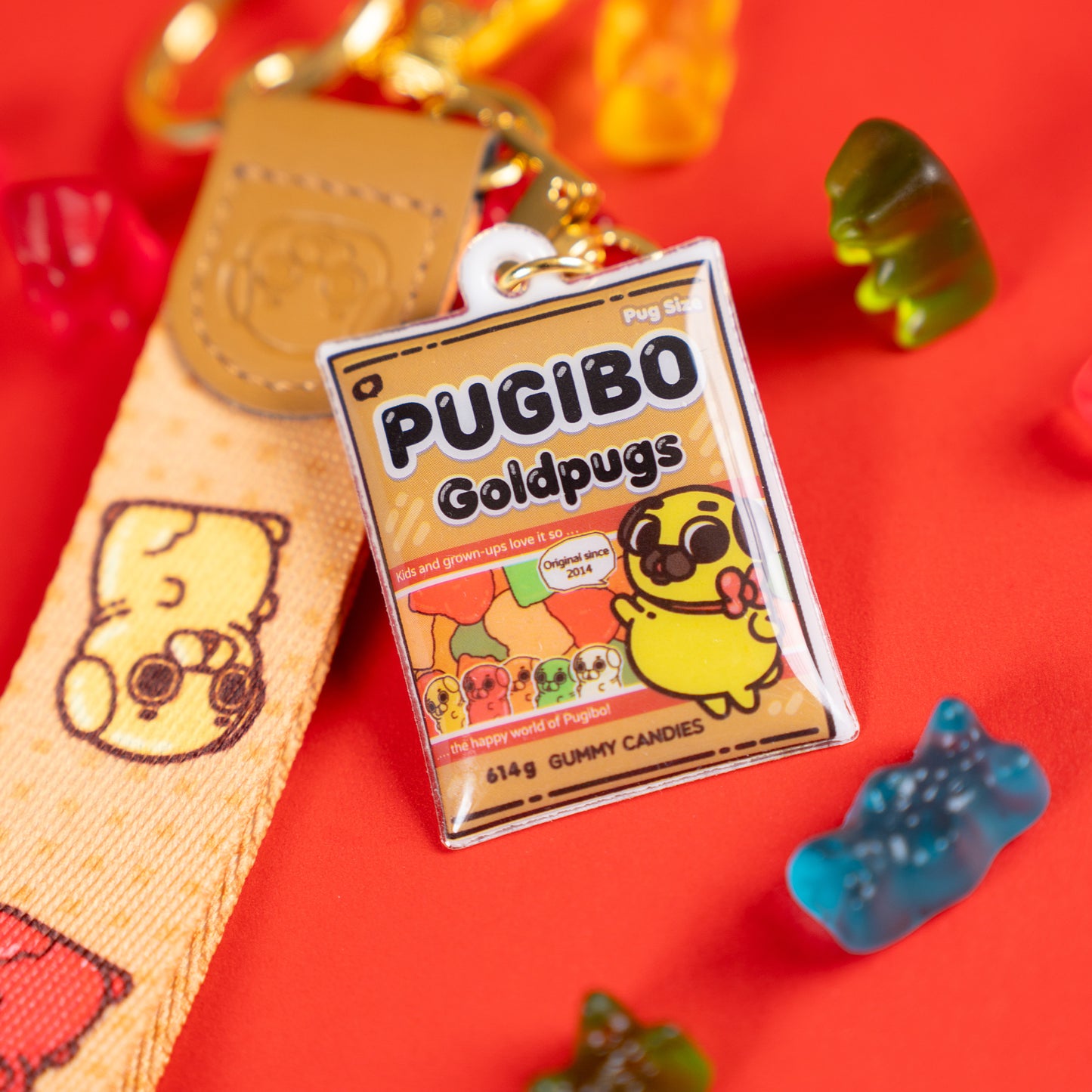 Keychain with a wrist strap featuring a design of candy-colored pugs (red and yellow pictured) with a leather-looking clasp embossed with a pug face. The leather-looking clasp is stitched to the wristlet and attached to a gold oval hoop, which has a gold jump ring, a gold lobster clasp, and a gold hook clasp attached with a cute charm featuring Puglie-themed bag of candies. They are designed in the style of "Haribo" and called "PUGIBO GOLDPUGS". The keychain is on a red background with gummy bear candies.