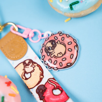 Keychain with a wrist strap featuring a design of various doughnuts with tiny pugs playing on them, with a leather-looking clasp embossed with a pug face. The leather-looking clasp is stitched to the wristlet and attached to a gold oval hoop. In focus is a pink hook clasp handing from the gold hoop with a cute charm featuring Puglie sitting in a pink doughnut with white sprinkles. The background is blue with doughnuts.